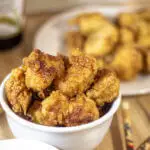 A bowl full of Korean fried chicken next to some Korean fried chicken sauce and chopsticks.