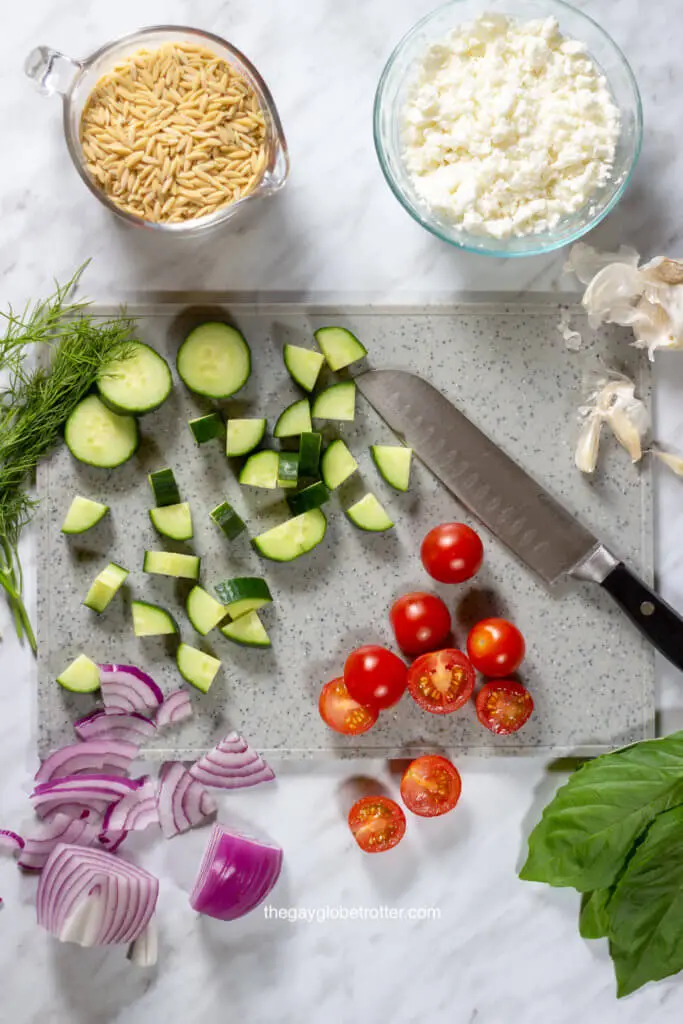 A cutting board filled with ingredients for orzo pasta salad like tomatoes, red onions, and cucumbers.