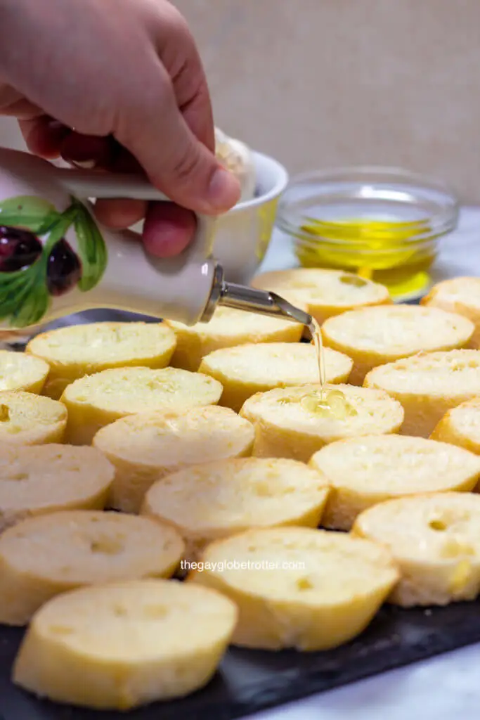 A hand pouring olive oil onto crostini slices.