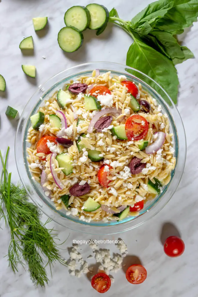 An overhead shot of Greek orzo pasta salad surrounded by dill, basil, cucumbers, and tomatoes.