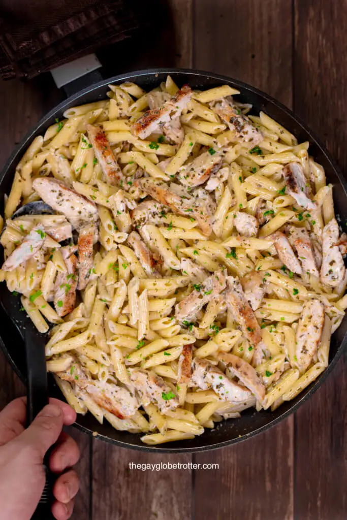 A hand scooping creamy pesto chicken pasta from a serving dish.