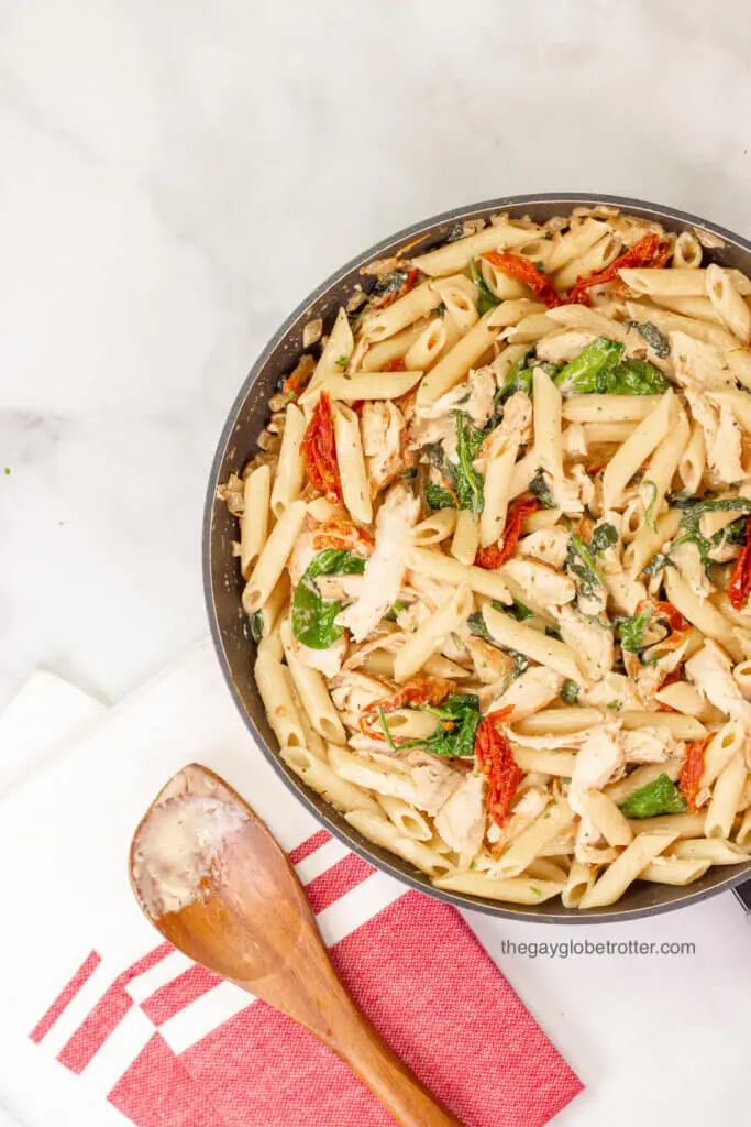 A pan of tuscan chicken pasta next to a wooden spoon that has just stirred the pasta.