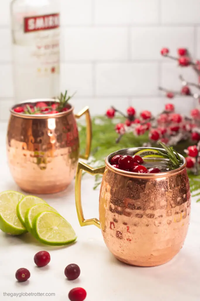 Two cranberry moscow mules next to a bottle of vodka.