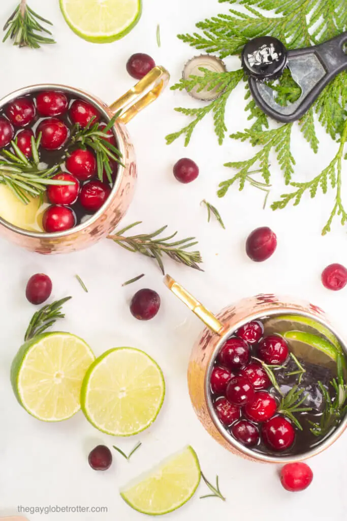 Two moscow mules with cranberries next to a bottle opener and limes.