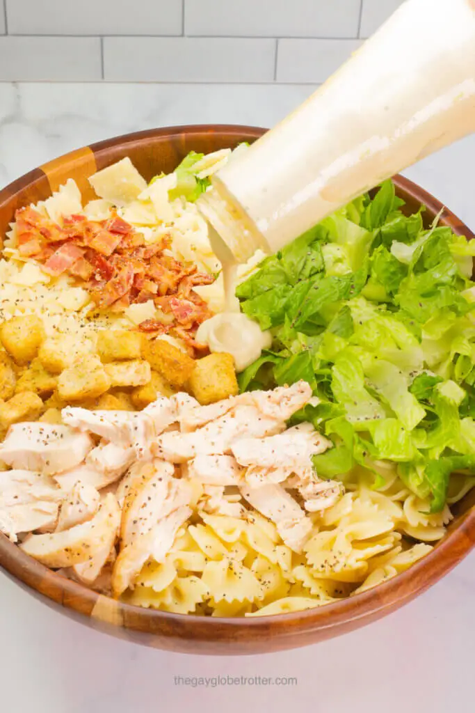 Caesar dressing being poured over caesar pasta salad ingredients. 