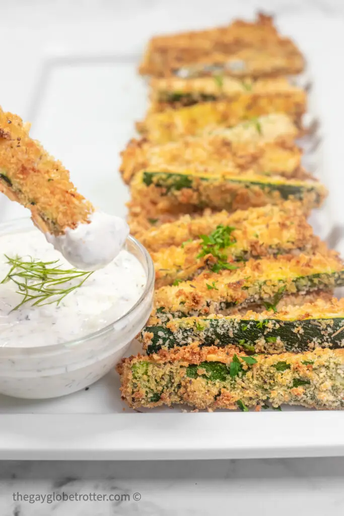 A breaded zucchini stick being dipped into creamy dill dip.