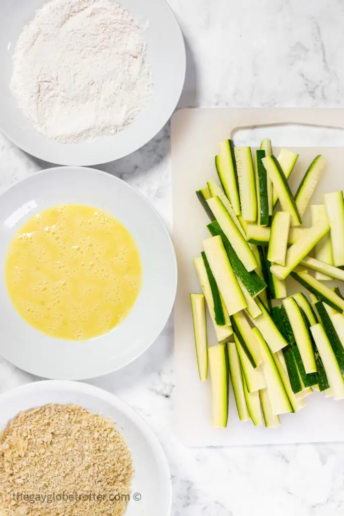 Zucchini sticks being breaded on a working surface.