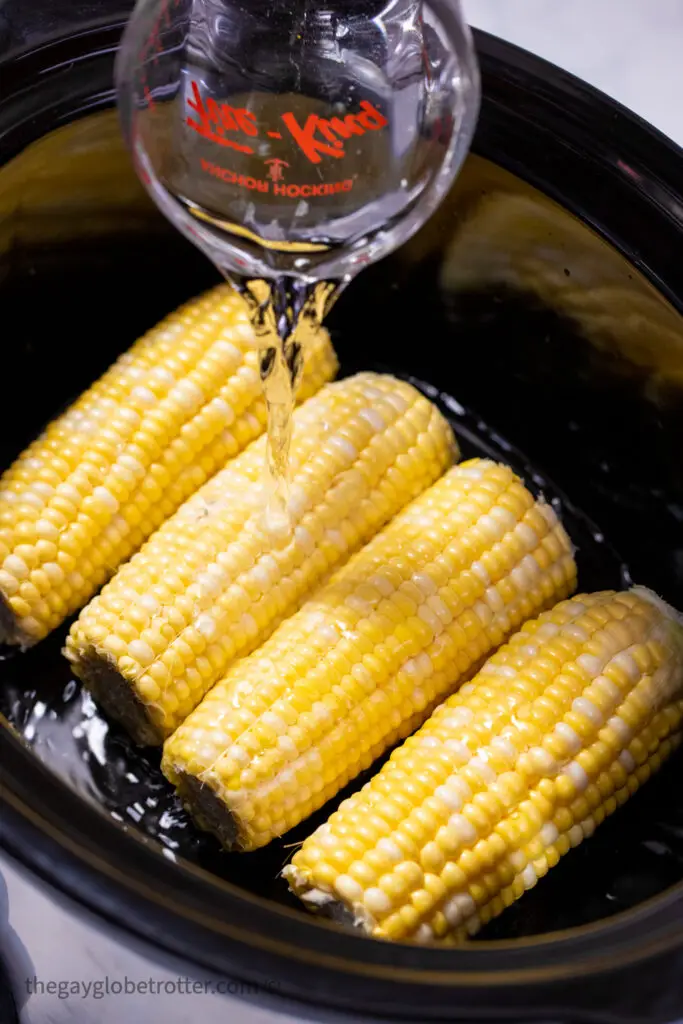 Corn arranged in a slow cooker while water is poured over it.