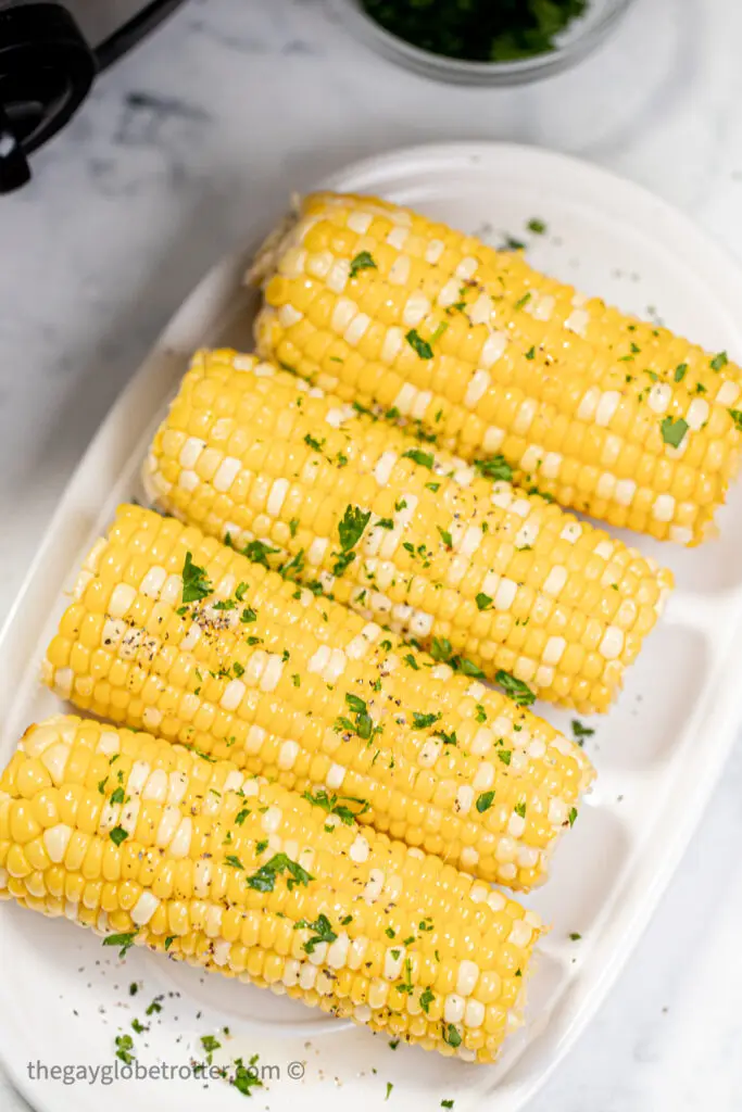 Corn in the cob on a platter garnished with parsley and butter.