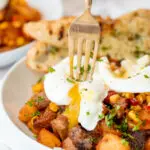 A fork breaking a poached egg on roast beef hash.